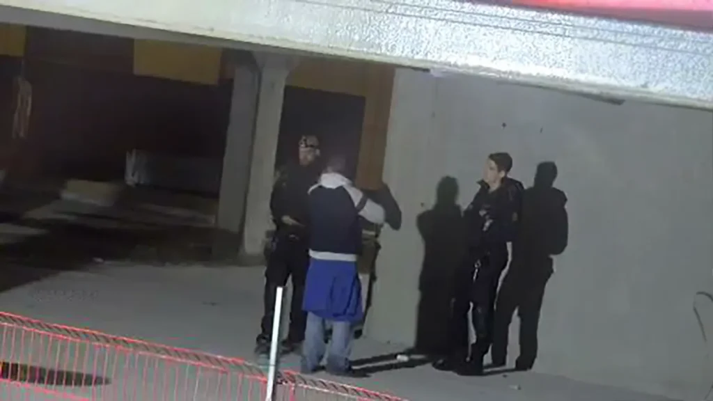 Politifolk avhører en mann utenfor en bygning om natten under sterkt lys med bemannet kameraovervåkning.